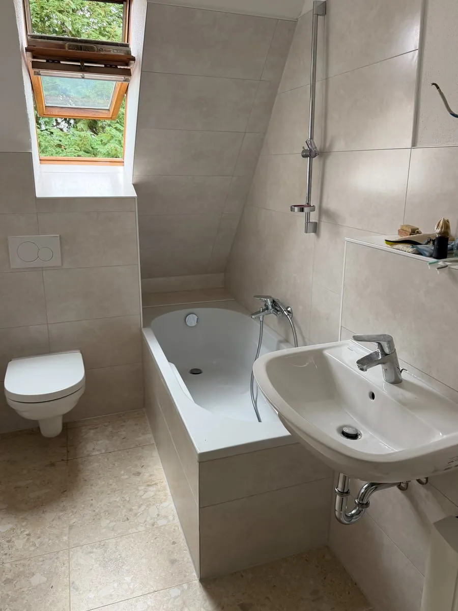 Dasselbe Dachbad nach der Sanierung: große helle Wand- und Bodenfliesen, gefliester Wannenrahmen, Wand-WC und Waschtisch