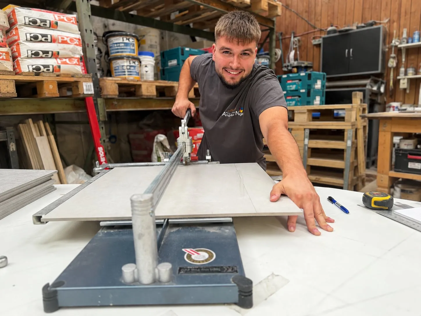 Junger Fliesenleger im Firmen-T-Shirt schneidet lächelnd eine großformatige Fliese an der Schneidemaschine in der Werkstatt