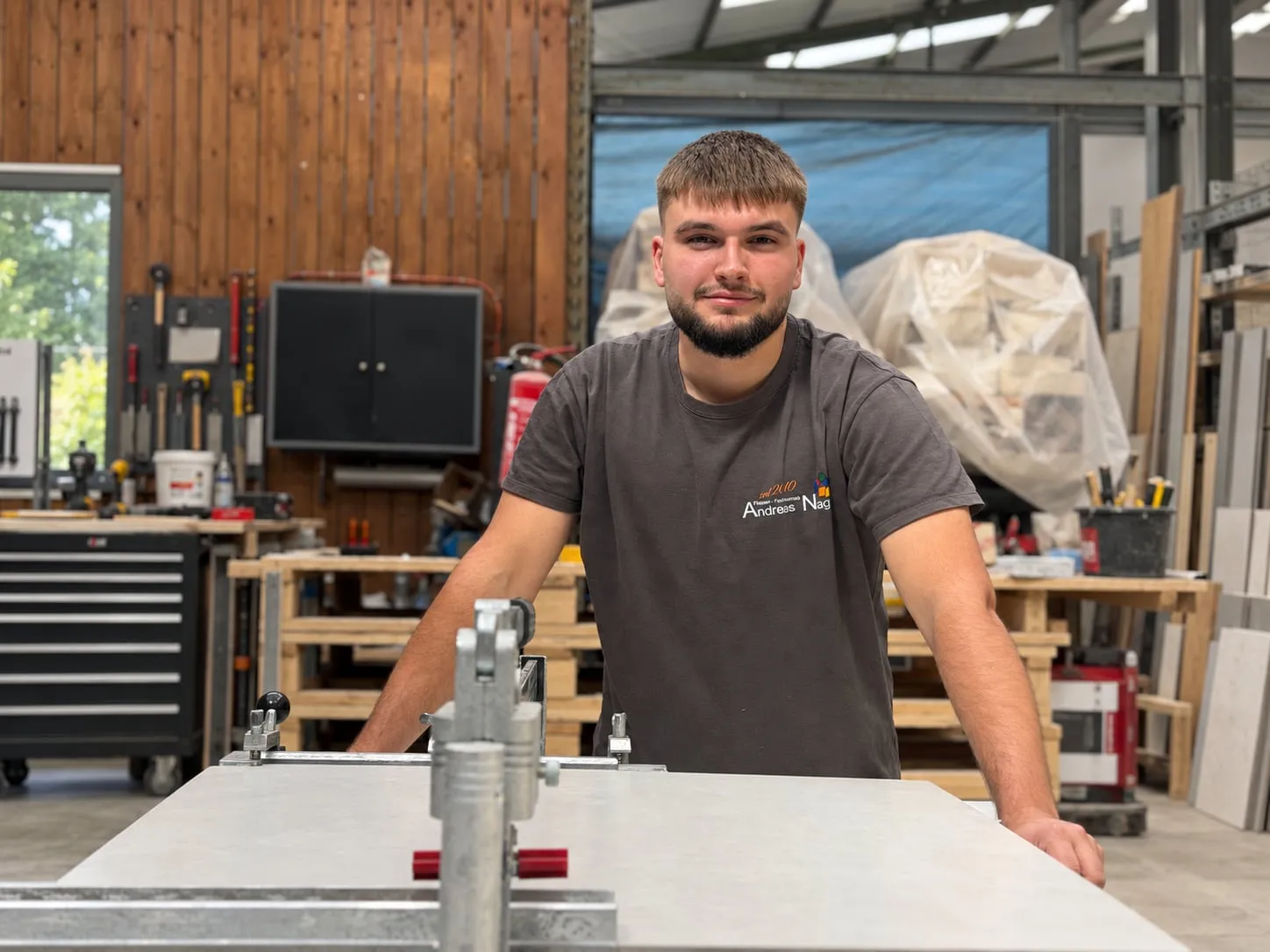 Junger Fliesenleger im Firmen-T-Shirt von Andreas Nagel stützt sich hinter der Schneidemaschine in der Werkstatt auf den Tisch
