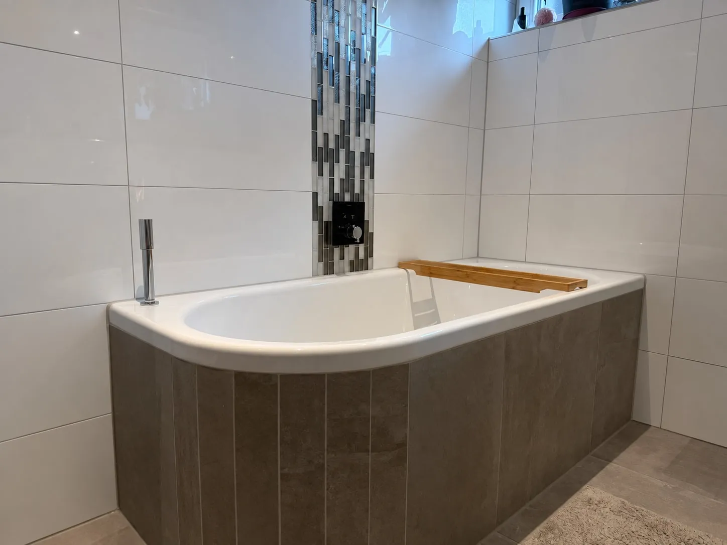 Badewanne mit Wannenverkleidung aus grauen Fliesen, dahinter ein senkrechtes Glasmosaikband in Grau- und Silbertönen an der weißen Wand