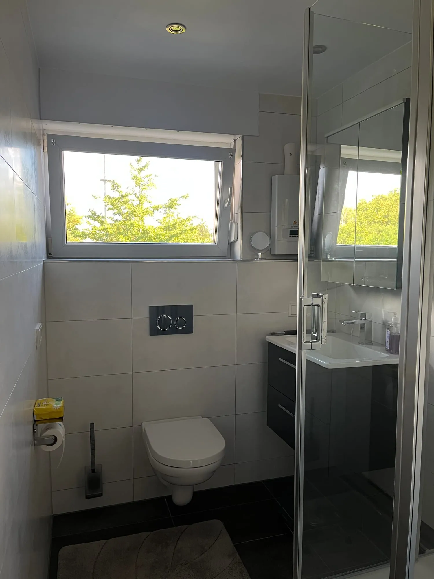 Helles Bad mit Wand-WC, Waschtisch und Glasdusche, graue Wandfliesen unter dem Fenster