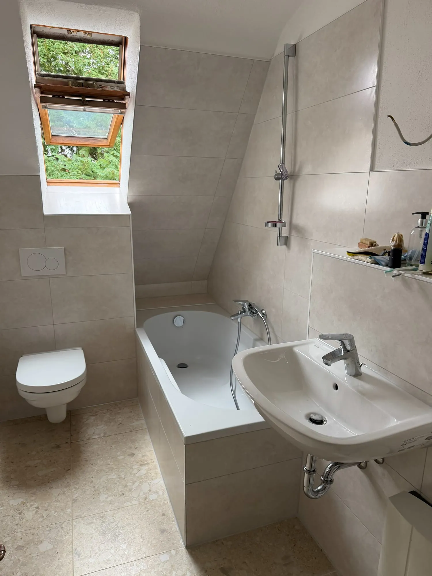 Bad unter der Dachschräge mit Dachfenster, eingefliester Badewanne und hellen Steinoptik-Fliesen