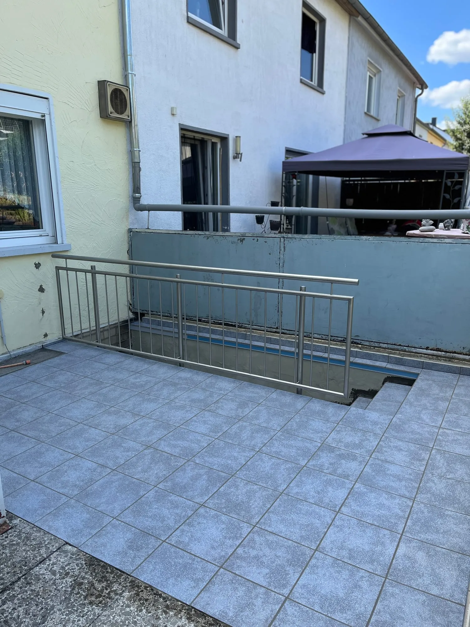 Terrasse mit grauen Fliesen und Kellerabgang, gesichert mit einem Edelstahlgeländer