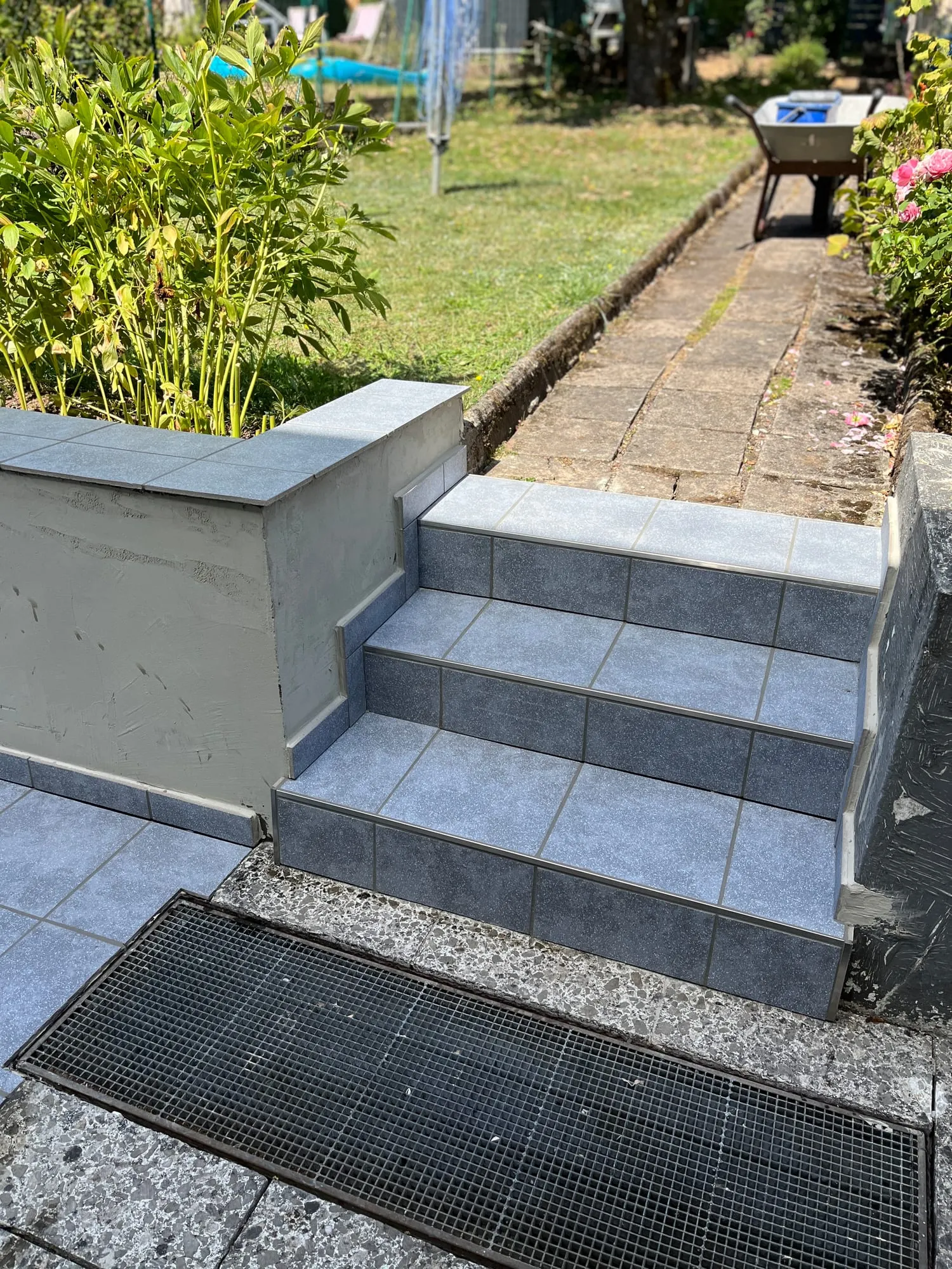 Kurze Außentreppe mit blaugrauen Platten, oben der Gartenweg, unten ein Gitterrost
