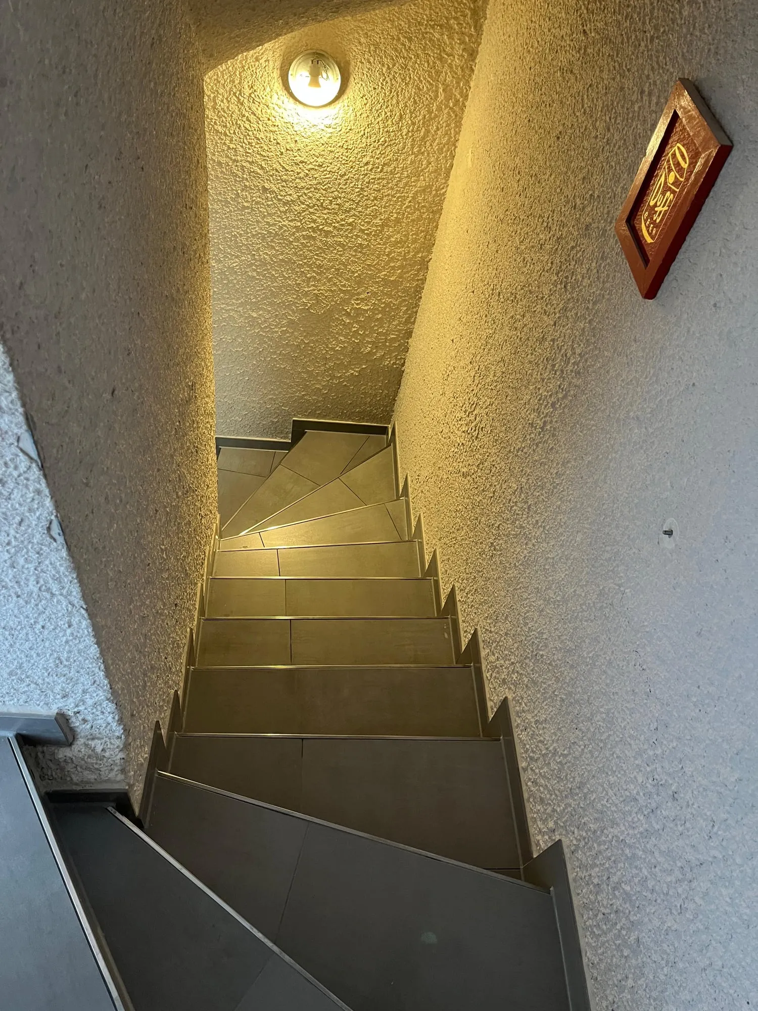 Blick von oben in ein schmales Treppenhaus mit dunkelgrau gefliesten Stufen und Winkelstufen am unteren Ende